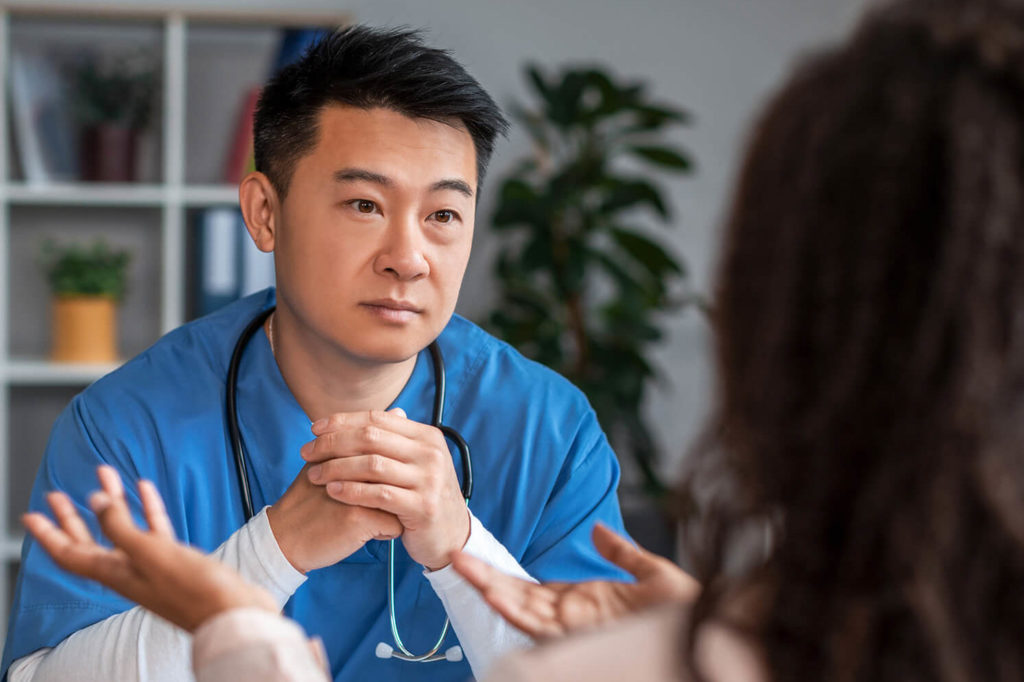 Serious millennial chinese guy doctor in uniform listens symptoms of african american lady patient in office
