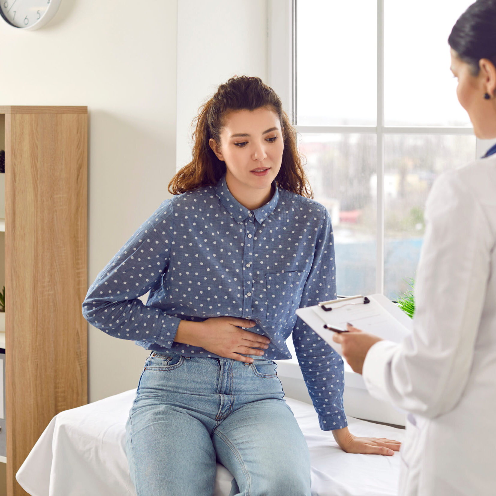 Female medical specialist consults young female patient who complains of abdominal pain. Woman experiences abdominal discomfort due to menstrual pain, pancreatitis, gastritis or diarrhea.