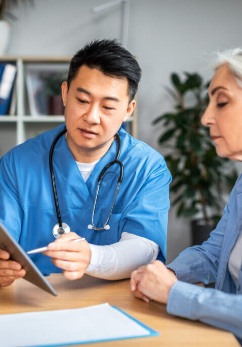 Serious middle aged asian man doctor showing tablet to retired european woman patient in clinic office interior. Life insurance, treatment with therapist, medical health care, receipt and service